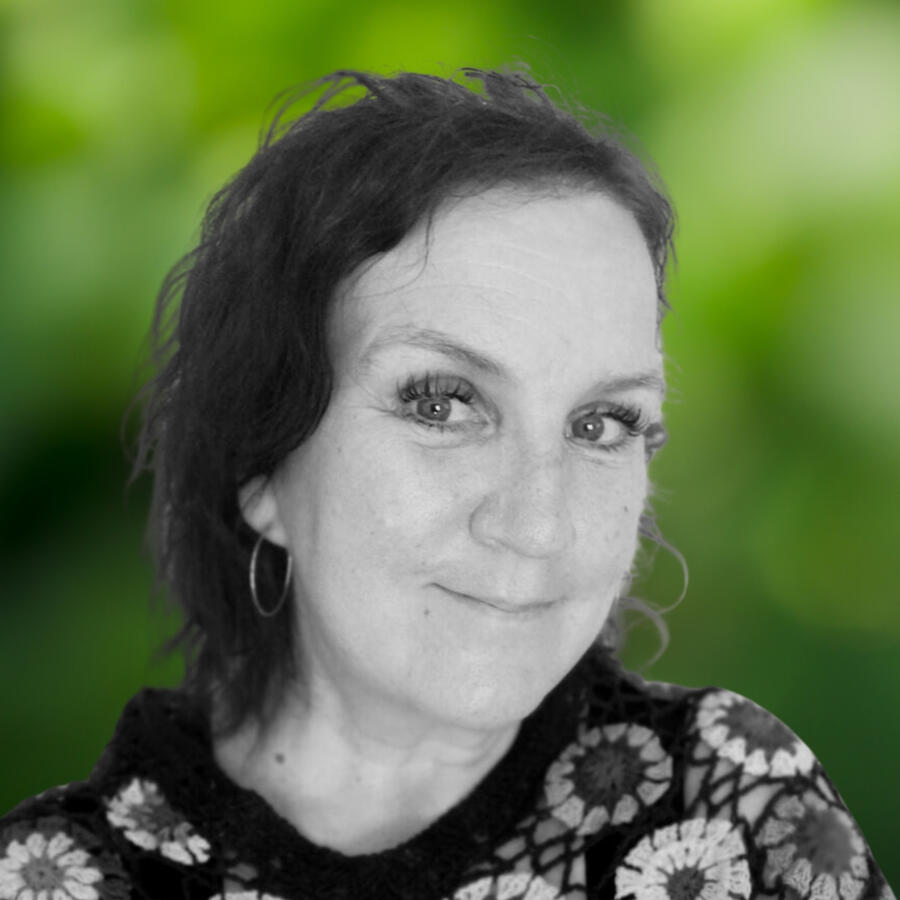 Nainen katsoo kameraan ja hymyilee. Hänellä on kiharat hiukset ja korvakorut.